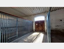 Innenansicht eines einfachen Stalls mit weißen Ziegelwänden, einer Holzeingangstür mit darüberliegendem Fenster und Metallgittern, die separate Boxen bilden. Der Boden besteht aus Pflastersteinen, und die Decke ist mit einer betonten Längsstruktur gestaltet.