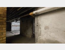 Ein alter, ungepflegter Kellerraum mit roh verputzten und teils unverkleideten Ziegelwänden. An der Decke verlaufen sichtbare Rohrleitungen, der Boden ist aus Beton, und es herrscht eine schummrige Beleuchtung.