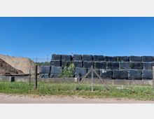 Ein Lagerplatz mit gestapelten, in schwarzer Folie verpackten Ballen, umgeben von einem Maschendrahtzaun. Im Hintergrund sind Erdhügel und ein blauer Himmel zu sehen.