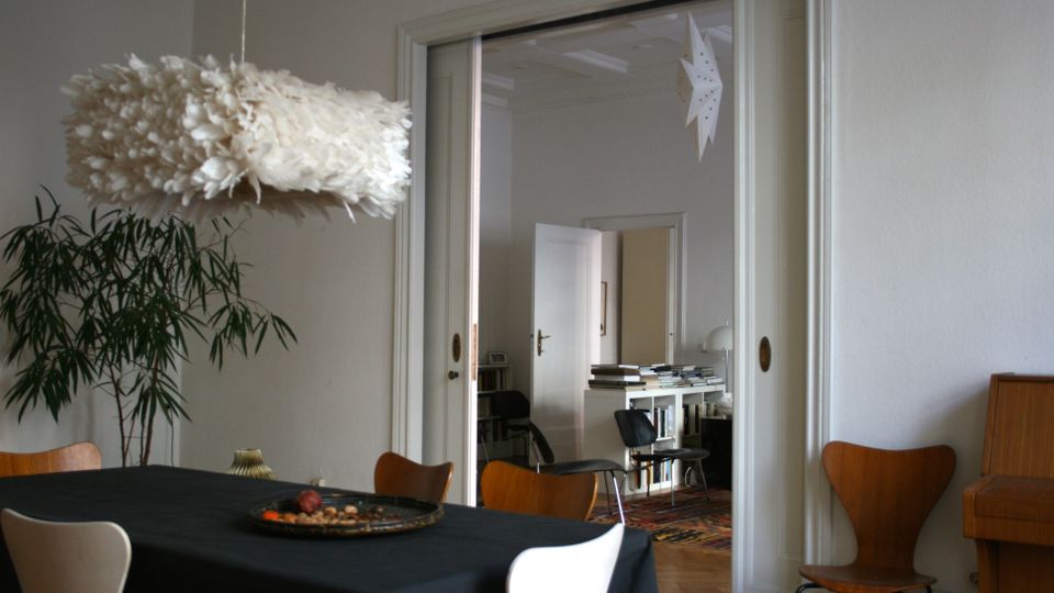 Heller Essbereich mit großem Tisch und Stühlen, einer federartigen Hängelampe und hohen weißen Wänden. Durch eine breite Schiebetüröffnung ist ein angrenzendes Zimmer mit Bücherstapel auf einem Regal, Teppichboden und geöffneten Türen zu sehen.