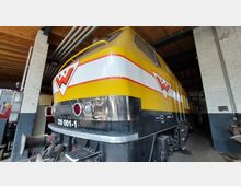 Gelbe Diesellokomotive mit roten und weißen Streifen in einer überdachten Halle, umgeben von Backsteinwänden und einem Holzdach. Auf der Front der Lok befindet sich ein "H.F.W."-Logo sowie die Kennzeichnung "320 001-1".