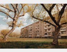 Ein mehrstöckiges Wohngebäude aus Beton mit Balkonen, umgeben von einer Wiese und großen Bäumen im Vordergrund. Im Hintergrund ist ein heller Himmel mit vereinzelten Wolken zu sehen.
