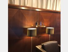 Hölzerne Wand mit geometrischem Rautenmuster und zwei angebrachten Leselampen. Vor der Wand stehen zwei Stehlampen mit gemusterten Schirmen und ein schwarzer Ledersessel.