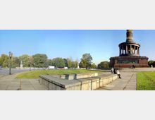 Die Abbildung zeigt die Siegessäule in Berlin, ein denkmalgeschützter Turm mit einer goldenen Statue an der Spitze, umgeben von einem runden Säulengang. Im Vordergrund befinden sich gepflasterte Wege, Steinbänke und grüne Rasenflächen; im Hintergrund stehen Bäume und eine Straße mit parkenden und fahrenden Autos.
