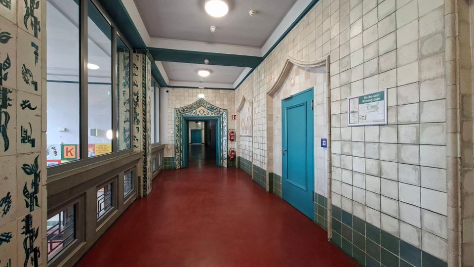 Langer Innenflur in einem Gebäude mit rotbraunem Boden, weiß gekachelten Wänden und runden Deckenleuchten. Rechts befindet sich eine türkisfarbene Tür, links große Fenster und dekorierte Türrahmen mit blaugrünen Ornamenten.