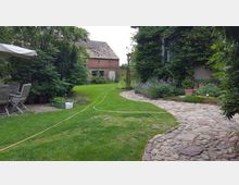 Ein gepflasterter Gartenweg führt durch einen grünen, gepflegten Garten mit Blumenbeeten und einem kleinen Rasenbereich. Im Hintergrund steht ein ziegelrotes Nebengebäude mit Schrägdach, umgeben von Bäumen und Sträuchern. Links befinden sich Gartenmöbel unter einem weißen Sonnenschirm.