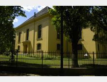 Gelbes, rechteckiges Gebäude mit Satteldach, flankiert von Bäumen und umgeben von einem schwarzen Gitterzaun. Vor dem Gebäude befindet sich eine grüne Rasenfläche, und im Hintergrund ist der Himmel teilweise bewölkt.