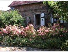 Backsteinbau mit Satteldach, grünen Fensterläden und einer offenen Tür, umgeben von einem üppigen Garten mit blühenden rosa Rosen und grünem Laub. Im Vordergrund befindet sich ein gepflasterter Weg.