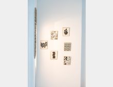 Eine weiße Wand mit sechs kleinen gerahmten Kunstwerken, die abstrakte Schwarz-Weiß-Muster und Blattformen zeigen, angeordnet in einer unregelmäßigen Formation. Links im Bild ist ein Teil einer Tür oder eines Rahmens sichtbar.