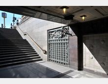 Treppenaufgang einer U-Bahn-Station mit einem Geländer auf der linken Seite, daneben eine verzierte Metalltür mit floralen Elementen. Die Umgebung besteht aus hellen Steinwänden und dekorativen Fliesen, beleuchtet durch Deckenlampen und Tageslicht.
