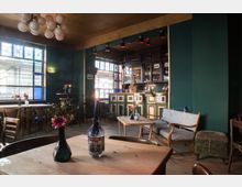 Gemütliche Bar mit dunklen grünen Wänden, Holzmöbeln und dekorativen Elementen wie Blumen und Kerzen auf den Tischen. Im Hintergrund befindet sich eine Theke mit Getränken und einem Regal, eingerahmt von großen Fenstern, die natürliches Licht hereinlassen.