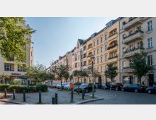 Straßenszene mit mehrstöckigen Wohnhäusern im Altbaustil, hellen Fassaden und Balkonen, gesäumt von Bäumen und parkenden Autos. Im Vordergrund befindet sich ein gepflasterter Gehweg mit Pollern, Straßenlaternen und einem Straßenschild mit der Aufschrift „Suarezstraße“.