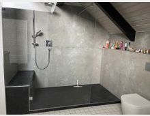 Modernes Badezimmer mit einer ebenerdigen Dusche, grauen Fliesenwänden und einer Glasabtrennung. Im Hintergrund befinden sich ein Regal mit Pflegeprodukten und eine schräg verlaufende Decke.