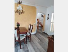 Essbereich in einem Wohnraum mit hellem Laminatboden, einem hölzernen Esstisch mit vier gepolsterten Stühlen, einer Messing-Deckenlampe und einer gelb gestrichenen Akzentwand. Im Hintergrund führen Holztreppen in ein anderes Stockwerk, und es sind Dekorationsgegenstände wie eine antike Wanduhr und Bilder an der Wand zu sehen.