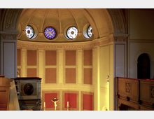 Innenraum einer Kirche mit gewölbter Decke, verziert mit Stuck- und Malereien. Oben sind vier runde, farbig gestaltete Kirchenfenster zu sehen, darunter roter und beiger Wandbereich mit einem Kruzifix in der Mitte, flankiert von Kerzen.