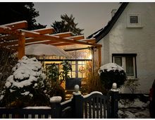 Einfamilienhaus mit weißer, verputzter Fassade und dunkel gerahmten Fenstern bei Nacht, umgeben von verschneiten Sträuchern und einem kleinen Garten. Der Eingang ist durch eine beleuchtete Glastür mit einem Türkranz sowie eine Holzpergola mit Schnee bedeckt gekennzeichnet.