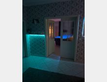 Innenraum mit gemusterten 3D-Wandpaneelen, hellem Fliesenboden und einer halb geöffneten Doppeltür mit Milchglas-Einsätzen. Links steht eine beleuchtete Theke mit türkisfarbenem LED-Licht; durch die Tür ist ein weiterer Raum mit blau beleuchtetem Billardtisch zu sehen.