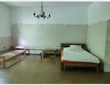 Ein einfacher Raum mit einer halbrenovierten Wand, die teils verputzt ist. Im Raum stehen zwei einzelne Betten, eines mit Matratze und Decke, das andere ohne Matratze, ein kleiner Couchtisch und ein einfacher Kronleuchter an der Decke.