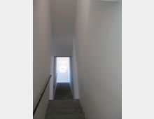 Treppenaufgang in einem schmalen, minimalistischen Innenraum mit weißen Wänden und einem dunkelgrauen Handlauf auf der linken Seite. Am oberen Ende der Treppe ist eine Wandlampe an der Rückwand zu sehen.