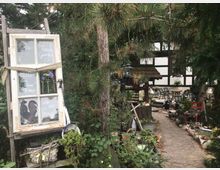 Ein rustikaler Garten mit verwinkeltem Steinweg, umgeben von dichten Nadelbäumen und dekorativen Elementen wie einem alten Fenster mit handgeschriebener Beschriftung. Im Hintergrund ist ein Fachwerkhaus und eine gepflasterte Terrasse mit Gartenmöbeln zu sehen.