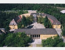 Luftaufnahme eines halbkreisförmig angelegten historischen Gebäudekomplexes mit gelber Fassade und grauen Dächern, umgeben von Bäumen und Grünflächen. Im Zentrum befindet sich ein gepflasterter Hof mit eingeschlossenem Hauptgebäude und großen Fenstern.