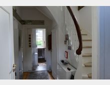 Heller Eingangsbereich eines Wohnhauses mit Holztreppe auf der rechten Seite, weißem Geländer und geschwungenem dunklen Holzhandlauf. Geradeaus führt ein Flur mit Glastür zu einem weiteren Raum, durch den Tageslicht hereinfällt.