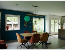 Esszimmer mit moderner Einrichtung, bestehend aus einem Holztisch und sechs braunen Stühlen, vor einer türkisfarbenen Wand mit großen Fenstern. Im Hintergrund sind Glastüren und ein Außenbereich mit grünem Garten sichtbar.