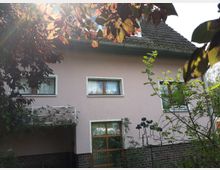 Zweistöckiges Haus mit rosa Fassade und braunem Ziegeldach, umgeben von Bäumen und Sträuchern. Im Vordergrund sind Fenster und ein kleiner Balkon mit Pflanzenbewuchs erkennbar.