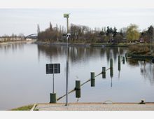 Flussansicht mit ruhigem Wasser, das von Uferböschungen und kahlen Bäumen gesäumt ist; im Vordergrund sind Pollerreihen im Wasser und eine gepflasterte Uferfläche mit Verkehrs- und Hinweisschildern sichtbar. Im Hintergrund führt eine Brücke über den Fluss.