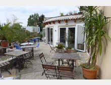 Außenbereich eines Cafés mit einer Terrasse aus Kopfsteinpflaster, umgeben von Pflanzen und Blumen in Töpfen. Es gibt Holztische mit Metallgestellen sowie Stühle, und die Terrasse grenzt an ein weißes Gebäude mit großen Glasfenstern und einem Dach aus Ziegeln.