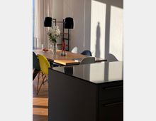 Ein moderner Essbereich mit einem Holztisch, umgeben von bunten Stühlen, zwei schwarzen Hängelampen und einer Vase mit weißen Blumen. Im Vordergrund eine schwarze Kücheninsel, im Hintergrund helle Vorhänge und ein Regal.