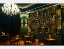 Ein gemütlicher Innenraum mit dunkelgrünen Wänden und Holzboden, dekoriert mit einem großen Kristallleuchter und einer Wand aus gestapelten Holzkisten, gefüllt mit Weinflaschen und Lichterketten. Im Vordergrund stehen Holztische und -stühle, auf den Tischen brennen Kerzen in Haltern.