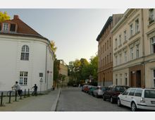 Straßenszene in einem Wohngebiet mit gepflasterter Straße, gesäumt von historischen Mehrfamilienhäusern mit verzierten Fassaden. Links steht ein rundes, weißes Gebäude mit schrägem Dach; geparkte Autos und Bäume sind im Hintergrund sichtbar.