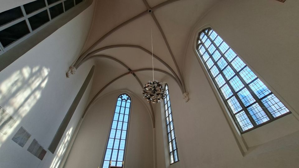 Innenraum einer Kirche mit weiß verputzten Wänden, hohem Kreuzrippengewölbe und großen, schmalen Spitzbogenfenstern mit Bleiverglasung. Von der Decke hängt ein moderner, kugelförmiger Kronleuchter.