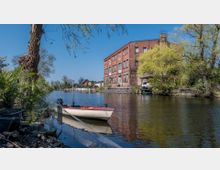 Backsteinbau an einem ruhigen Fluss, flankiert von Bäumen und Vegetation. Im Vordergrund liegt ein kleines weiß-rotes Boot am Ufer, während sich im Hintergrund Häuser und Gärten entlang des Wassers befinden.