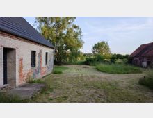 Ein ländliches Anwesen mit einem verputzten Ziegelgebäude links und einem weiteren Gebäude mit Ziegeldach rechts, umgeben von einer Wiese, Bäumen und hohem Gras. Im Hintergrund erstreckt sich eine offene Landschaft unter klarem Himmel.