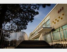 Ein modernes Gebäude mit großer Fensterfront und einem Balkon; an der Fassade ist der Schriftzug „BAR“ zu sehen. Im Vordergrund führen geschwungene Metallgeländer eine Rampe hinauf, und rechts ist ein auskragendes Vordach sichtbar, während im Hintergrund Bäume und blauer Himmel zu erkennen sind.