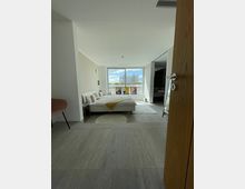 Blick in ein modernes Schlafzimmer mit großen Fenstern, die eine Ansicht auf ein Gebäude und den Himmel bieten. Der Raum ist hell eingerichtet, mit einem Doppelbett, beige-gelben Decken und klaren Linien; auf dem Boden liegt ein kleiner runder Teppich.