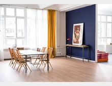 Heller Raum mit großen weißen Sprossenfenstern, einem Esstisch aus Metall mit sechs Holzstühlen und gelben Vorhängen. Im Hintergrund steht ein schmaler Konsolentisch vor einer dunkelblauen Wand, darauf befindet sich ein gerahmtes Kunstwerk und eine rote Tischlampe.