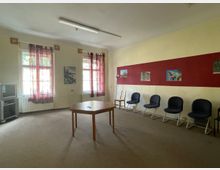 Ein einfacher Gemeinschaftsraum mit beigefarbenen Wänden, einer roten Akzentwand, an der mehrere Bilder hängen, und zwei Fenstern mit durchscheinenden, violetten Vorhängen. Der Raum enthält eine Reihe blauer Stühle, einen Holztisch in der Mitte, einen Fernseher in der Ecke und eine Stehlampe.