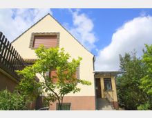 Zweistöckiges Wohnhaus mit gelber Fassade, braunen Fensterläden und einer Außentreppe, die zum Eingang führt. Im Vordergrund stehen ein Baum und Sträucher, links begrenzt ein Holzzaun den Bereich, bei klarem Himmel.