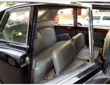 Innenraum eines klassischen Autos mit geöffneter Tür, ausgestattet mit grauen Ledersitzen und Holzelementen an den Türen. Auf der Rückbank liegt eine weiße Rose, und im Hintergrund sind Autos und eine Häuserfront auf einer Straße zu sehen.