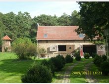 Backsteingebäude mit roten Dachziegeln, vermutlich ein Stallgebäude, umgeben von gepflegtem Rasen, Sträuchern und Bäumen. Im Hintergrund ist ein bewaldeter Bereich zu sehen, links im Bild ein kleiner Turm mit ähnlicher Dachkonstruktion.