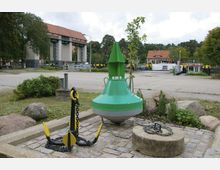 Grüne Boje und schwarz-gelber Anker ausgestellt auf gepflasterter Fläche, umgeben von Grasflächen und Büschen. Im Hintergrund ein Hafengebäude mit rotem Dach, ein Kanal mit Booten und Bäume.