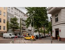 Straßenszene in einer Wohngegend mit mehrstöckigen Altbauhäusern, Bäumen entlang der Gehwege und parkenden Autos. Auf einem Straßenschild sind die Straßennamen erkennbar, und im Hintergrund sind kleine Geschäfte sowie ein gelber Briefkasten zu sehen.