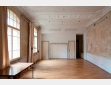 Großer, leerer Innenraum mit hellem Holzfußboden, hohen Fenstern mit Vorhängen an der linken Wand und einer teils abgenutzten Putzwand rechts. Die Decke ist hoch und reich verziert mit Stuckfeldern; an der Rückwand befinden sich zwei Türen, ein offener Durchgang und ein freistehender Kleiderständer.