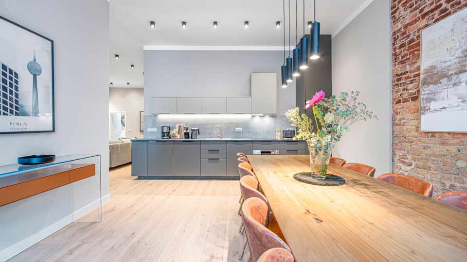 Offener Wohn- und Essbereich mit moderner grauer Einbauküche, langem Holzesstisch und mehreren Polsterstühlen unter schwarzen Pendelleuchten. Helle Holzböden, eingelassene Deckenleuchten, eine freiliegende Ziegelwand und gerahmte Wandbilder prägen den Raum.