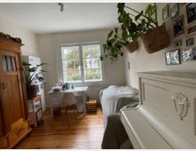 Helles Schlafzimmer mit Holzdielen, einem Bett an der rechten Wand und einem großen Fenster am Ende des Raums mit Schreibtisch davor. Links stehen ein Holzschrank und ein Kühlschrank, rechts hängen Pflanzenkörbe und mehrere gerahmte Fotos an der Wand.