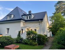 Einfamilienhaus mit weißer Fassade und dunkelgrauem Schrägdach, umgeben von einem gepflegten Garten mit Büschen, Bäumen und einem gepflasterten Weg. Links steht eine weiße Laterne, und im Hintergrund ist blauer Himmel sichtbar.