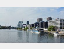 Stadtansicht von Berlin entlang der Spree mit modernen Bürogebäuden, darunter das Zalando-Gebäude, und einem anliegenden Hausboot. Im Hintergrund sind der Berliner Fernsehturm, Baukräne und ältere Industriebauten zu sehen.
